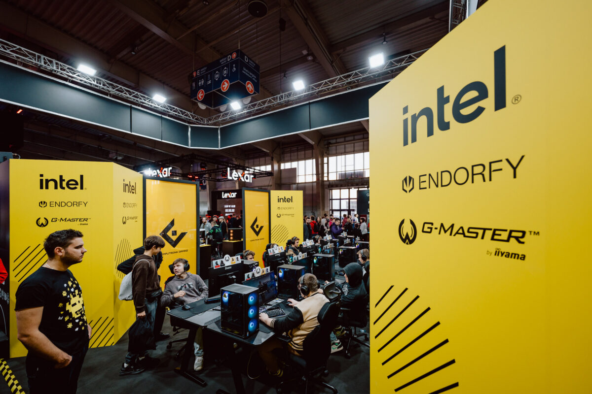 Ein Gaming-Event mit Menschen, die an mehreren Ständen Computer spielen, mit großen gelben Bannern mit den Logos von Intel, Endorfy und G-MASTER von iiyama. Im Hintergrund sind Zuschauer und Teilnehmer zu sehen. 