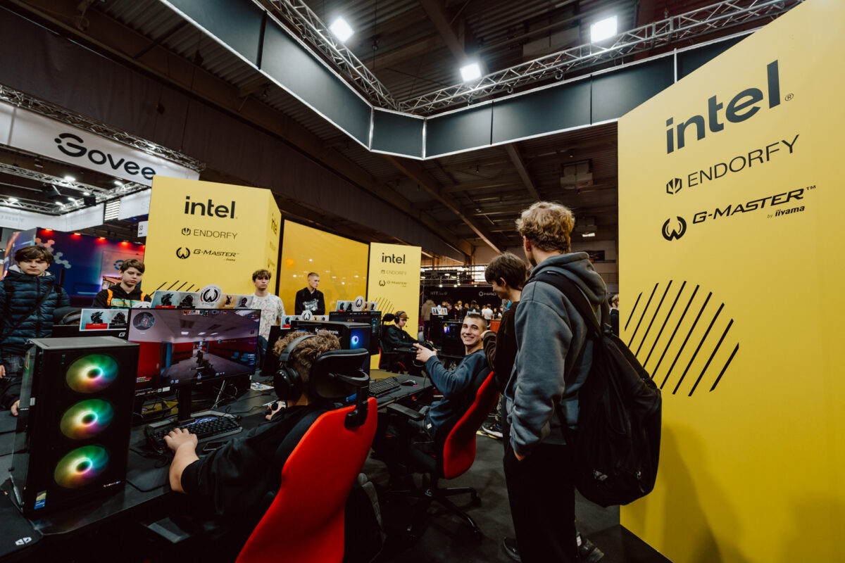 Eine Gruppe junger Leute versammelt sich an einem von Intel gesponserten Stand auf einer Messe um Spielecomputer mit gelben Bannern, auf denen die Logos von Intel, Endorfa und G-Master abgebildet sind. Einige sitzen und spielen, während andere stehen und zusehen. 
