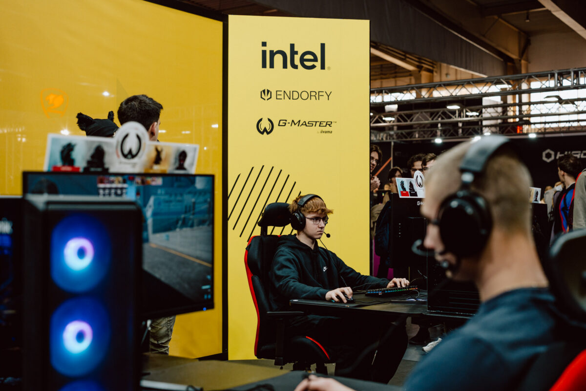 Ein junger Mann sitzt bei einem Gaming-Event vor einem großen gelben Intel-Display an einem Gaming-Computer, während andere Gamer in der Nähe mit Headsets gegeneinander antreten.