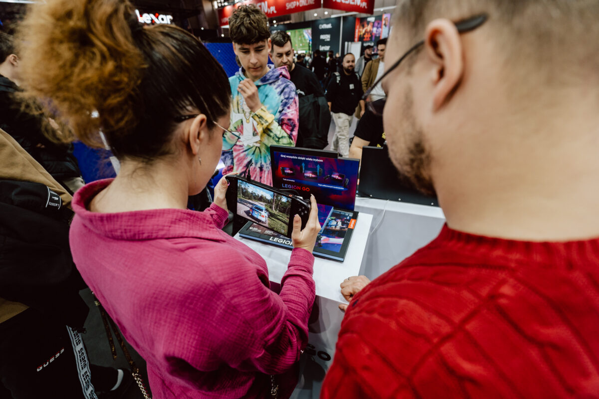 Eine Frau und ein Mann, beide in roten Blusen, sehen sich auf einer Technologiemesse Gaming-Laptops an. Ein Jugendlicher und ein weiterer Mann stehen in der Nähe, im Hintergrund sind Stände und elektronische Displays zu sehen. 