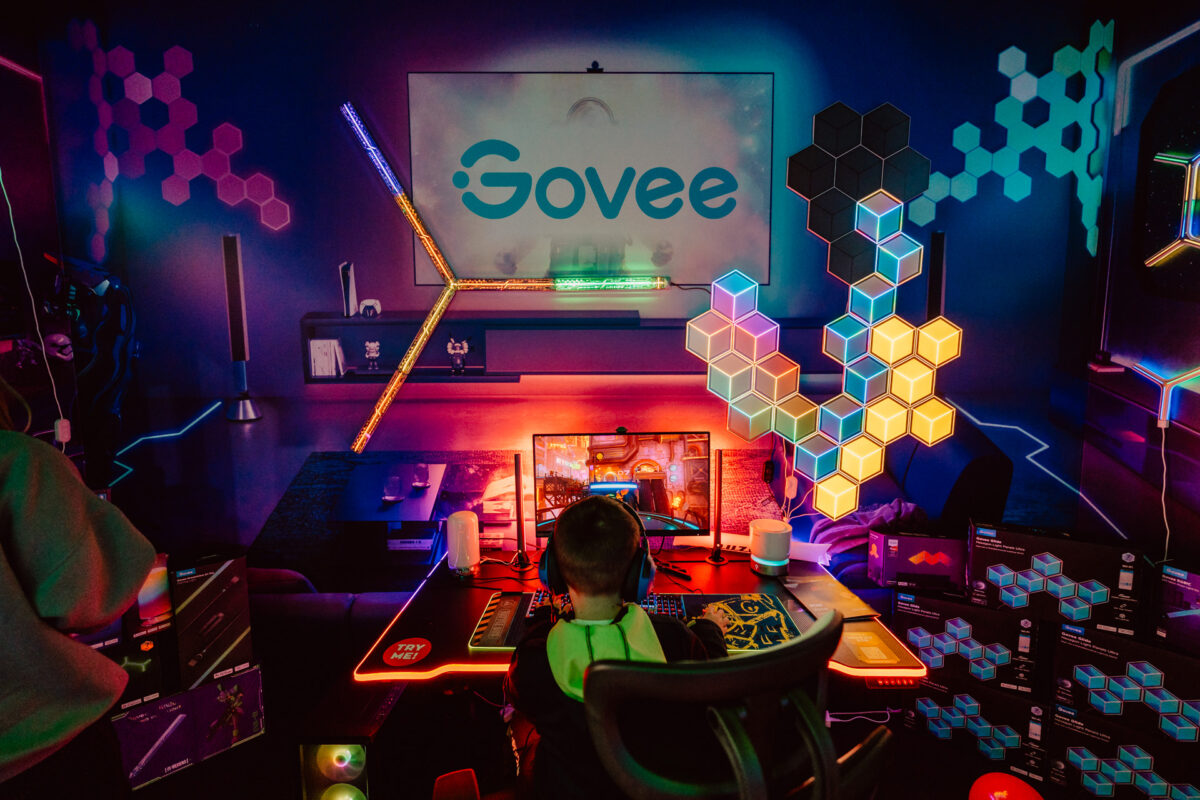 Eine Person sitzt an einem Spieltisch mit farbigen LED-Lichtern, sechseckigen Wandpaneelen und mehreren Monitoren und blickt auf einen großen Bildschirm mit dem 'Govee'-Logo in einem lebendigen, futuristischen Raum.