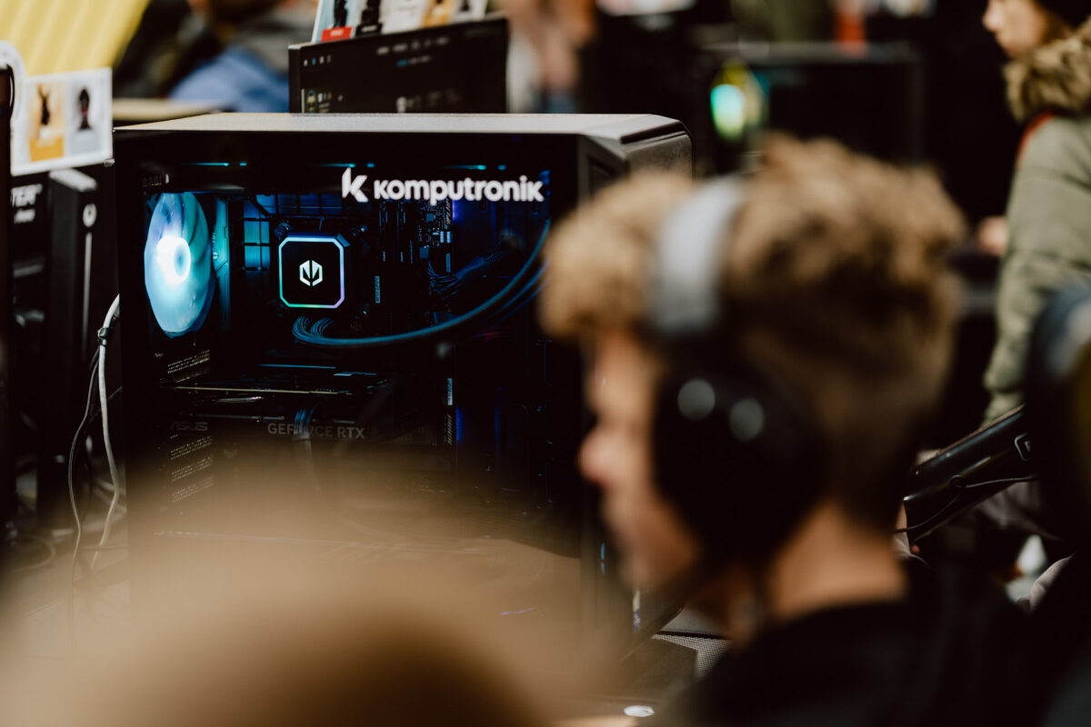 Eine Person mit Kopfhörern sitzt vor einem Gaming-Desktop-Computer mit LED-Beleuchtung und dem Komputronik-Logo auf dem Gehäuse, mit verschwommenen Menschen und Computerbildschirmen im Hintergrund.