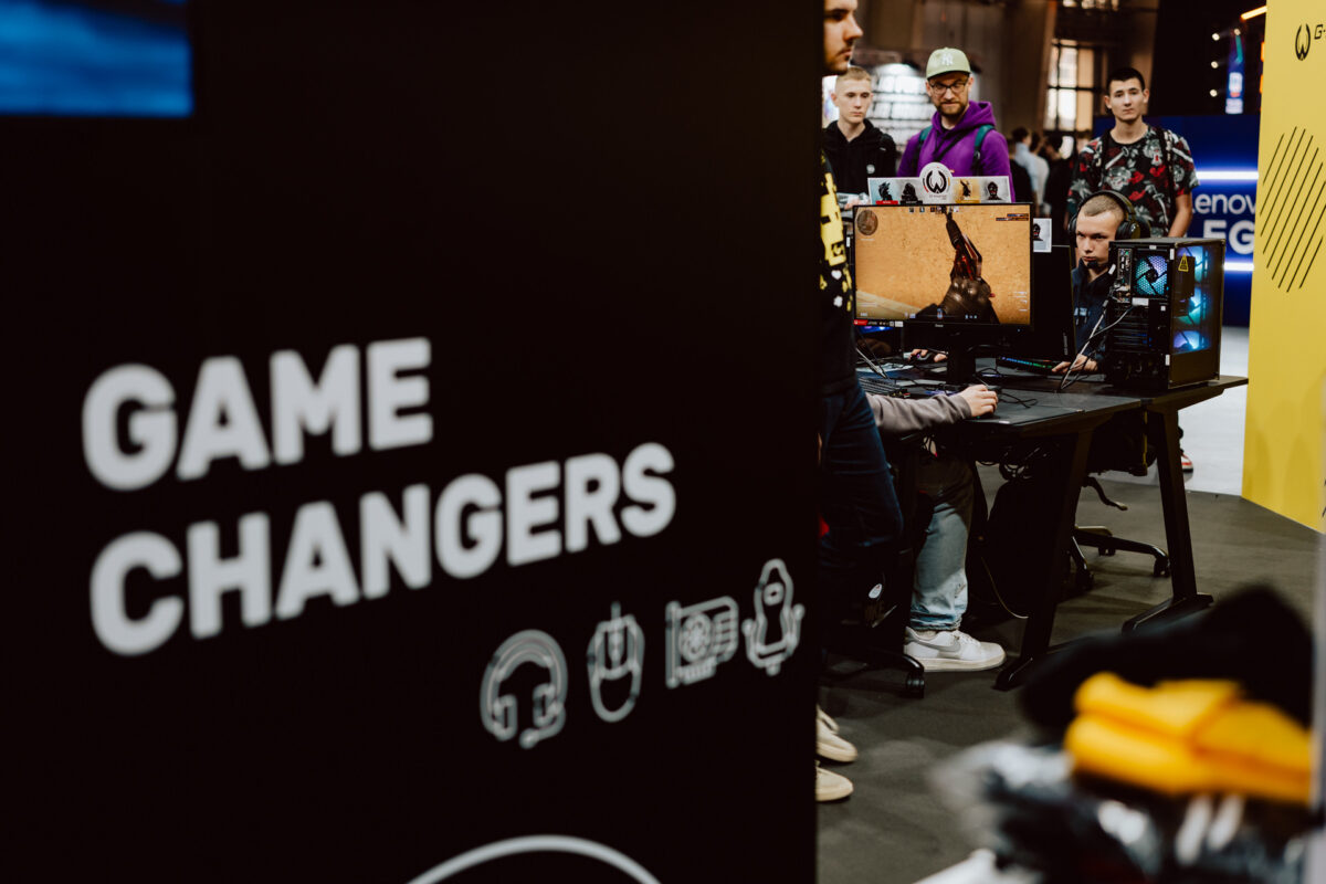 Eine Gruppe von Leuten bei einem Gaming-Event beobachtet jemanden, der einen Ego-Shooter auf einem Computer spielt. Im Vordergrund steht ein großes schwarzes Schild mit der Aufschrift 'GAME CHANGERS' und Spielsymbolen. 