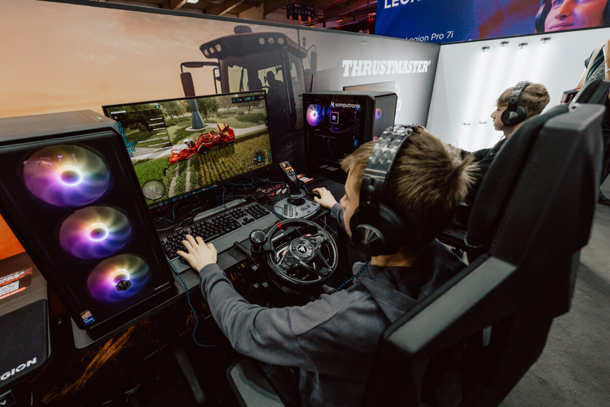 Zwei Jungen mit Kopfhörern spielen auf einem Gaming-Event oder einer Convention Landwirtschaftssimulationen auf High-End-Computern mit RGB-Beleuchtung, Lenkrädern und Joystick-Controllern.
