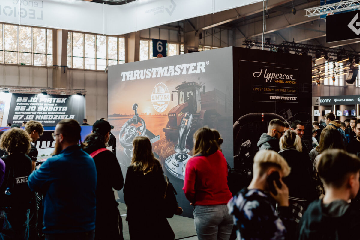 Eine Menschenmenge versammelt sich bei einer Indoor-Veranstaltung um einen großen Thrustmaster-Stand, an dem Rennsimulationsgeräte und Werbedisplays ausgestellt sind. Im Hintergrund sind helle Lichter und andere Stände zu sehen. 