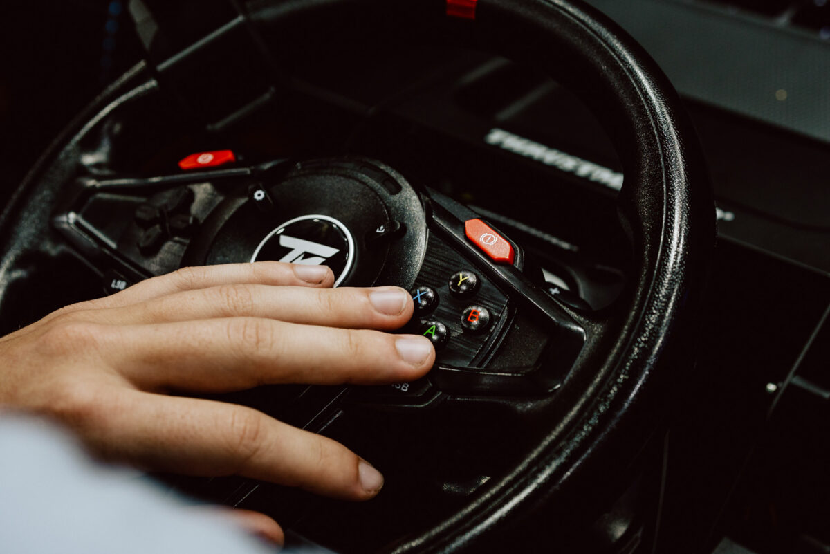 Nahaufnahme der Hand einer Person, die die Tasten eines schwarzen Rennrad-Controllers mit verschiedenen farbigen Tasten und einem Logo in der Mitte drückt. Die Szene erinnert an ein Videospiel oder einen Rennsimulator. 