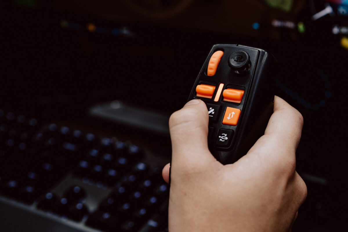 Eine Person, die einen schwarz-orangenen Joystick-Controller vor einer unscharfen Tastatur und einem Computermonitor hält, wahrscheinlich in einem Spiel oder einer Simulation beschäftigt.