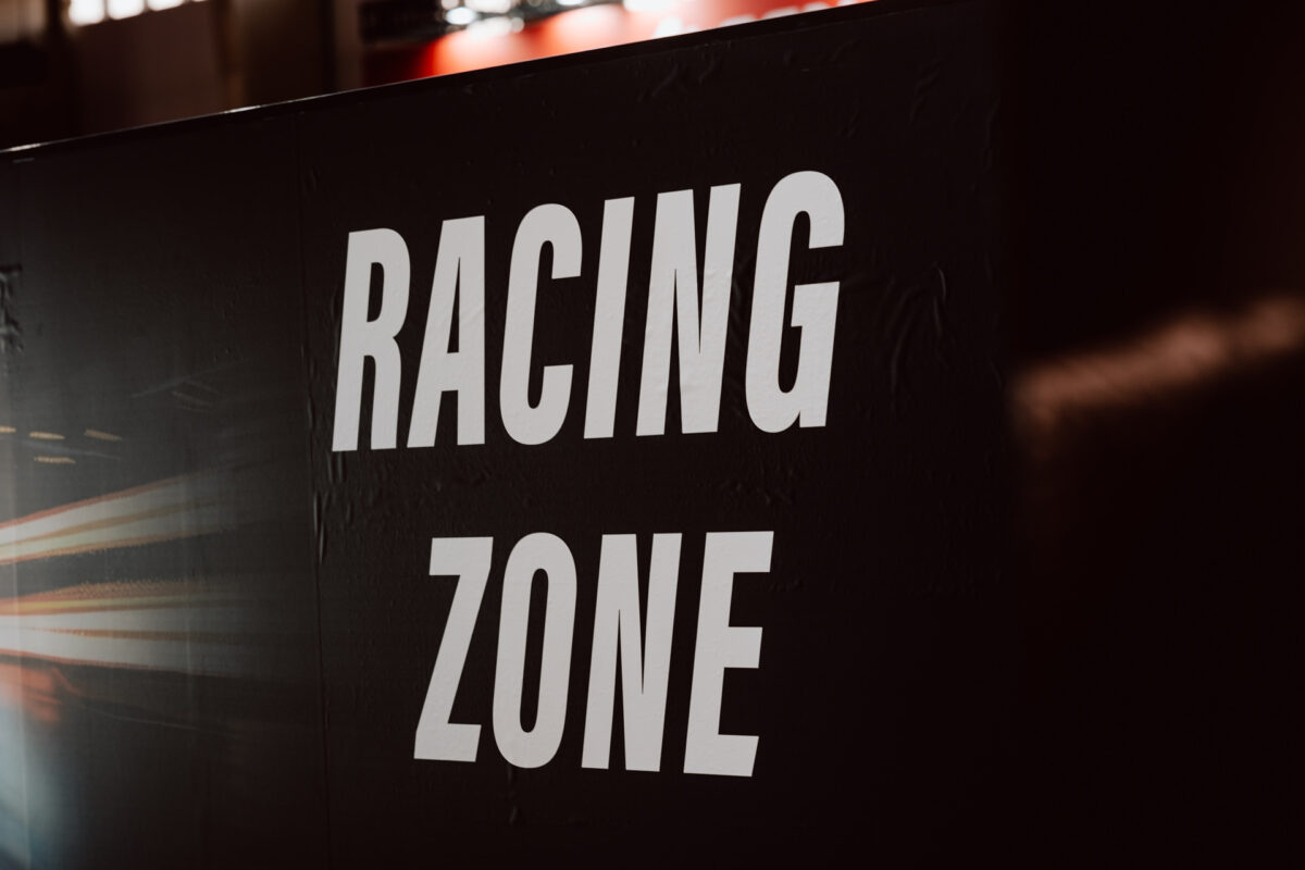 Eine schwarze Wand mit einem großen weißen Schriftzug 'RACING ZONE', an der links unscharfe Lichter zu sehen sind, die Bewegung oder Geschwindigkeit suggerieren.