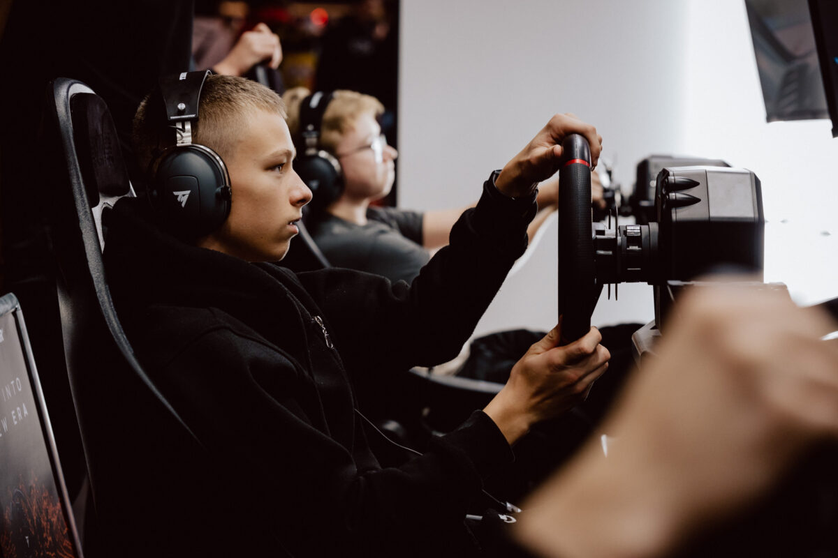 Ein Junge mit Kopfhörern hält ein Rennrad in der Hand und konzentriert sich auf einen Fahrsimulator. Eine andere Person im Hintergrund spielt ebenfalls, beide sitzen an Spielkonsolen. 