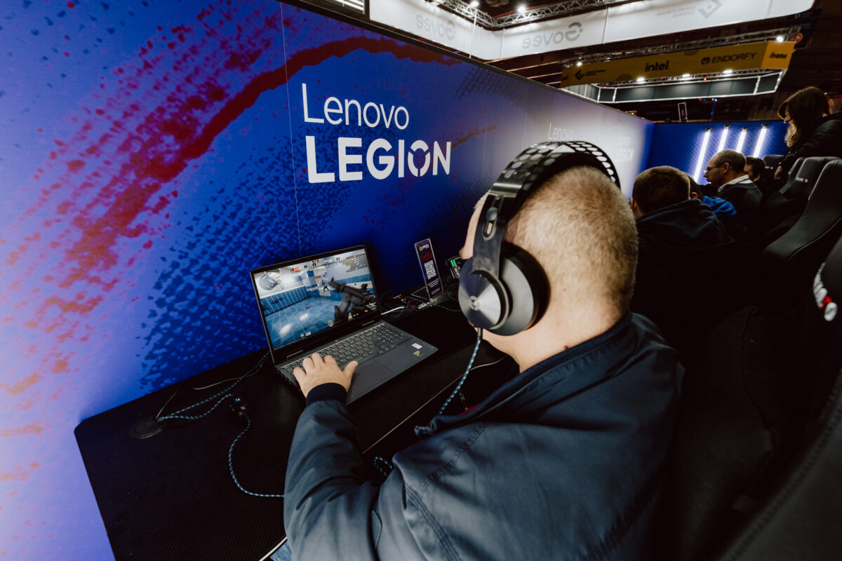 Die Person, die die Kopfhörer trägt, spielt ein Videospiel auf einem Laptop am Lenovo Legion Stand, umgeben von blauen Markenwänden, zusammen mit anderen Teilnehmern des Gaming-Events.