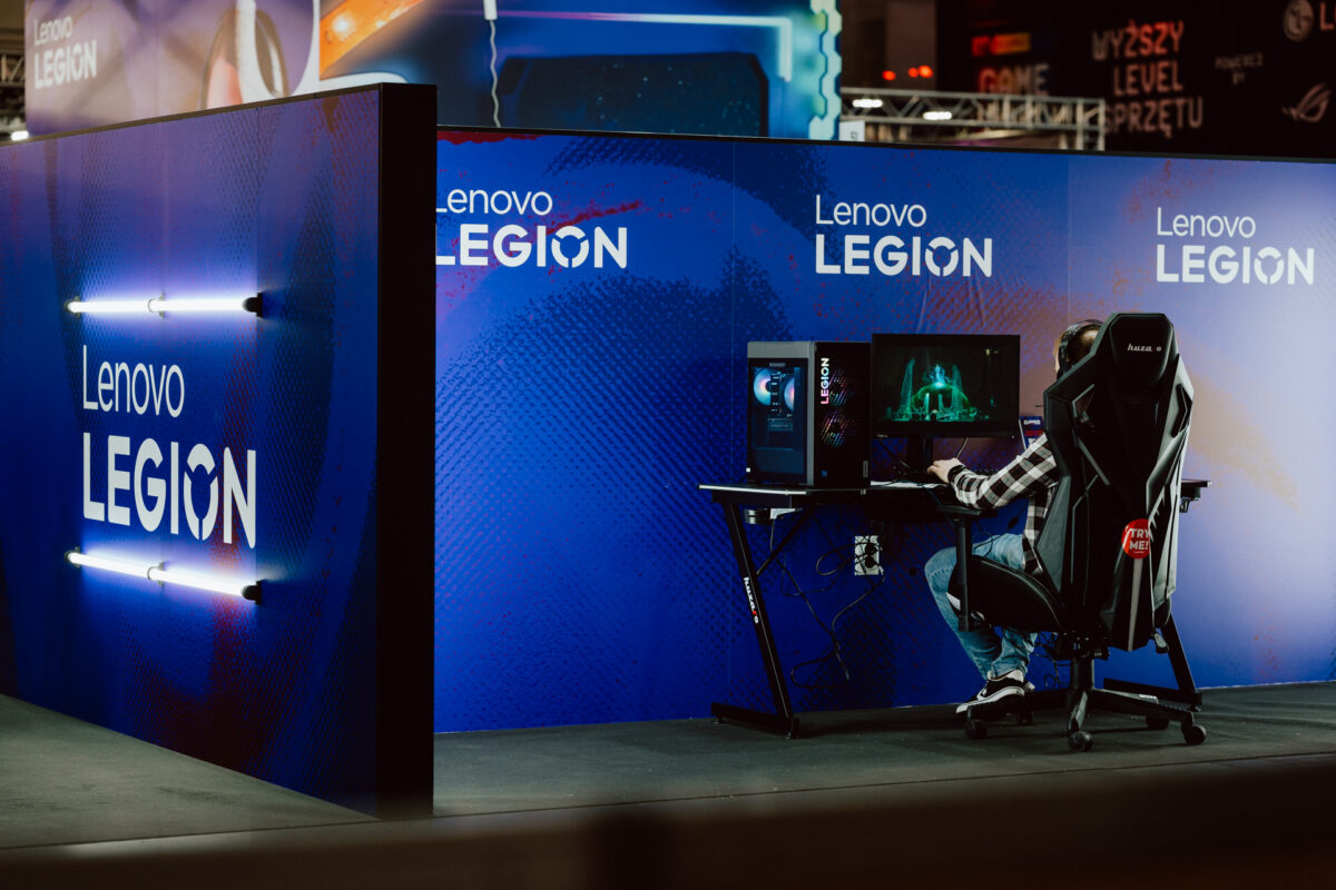 Eine Person sitzt in einem Gaming-Stuhl an einem Schreibtisch mit einem Computer-Set in einem Stand mit blauem Lenovo Legion Branding auf einer Gaming-Veranstaltung. Neonlichter beleuchten die Seitenwand des Standes. 