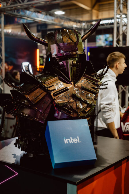 Eine detaillierte, dunkle Samurai-Rüstung aus Metall steht auf einem Tisch mit einer blauen Intel-Box davor. Im Hintergrund sind Menschen und Technologiestände zu sehen. 
