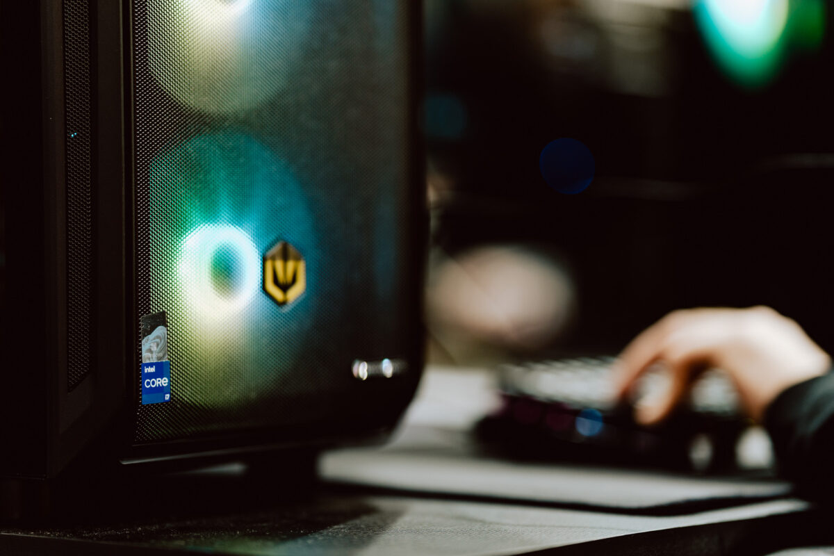 Nahaufnahme eines Gaming-Computer-Towers mit farbigen LED-Lichtern und einem sichtbaren Intel Core-Aufkleber, daneben eine verschwommene Hand, die auf einer Tastatur im Hintergrund tippt.
