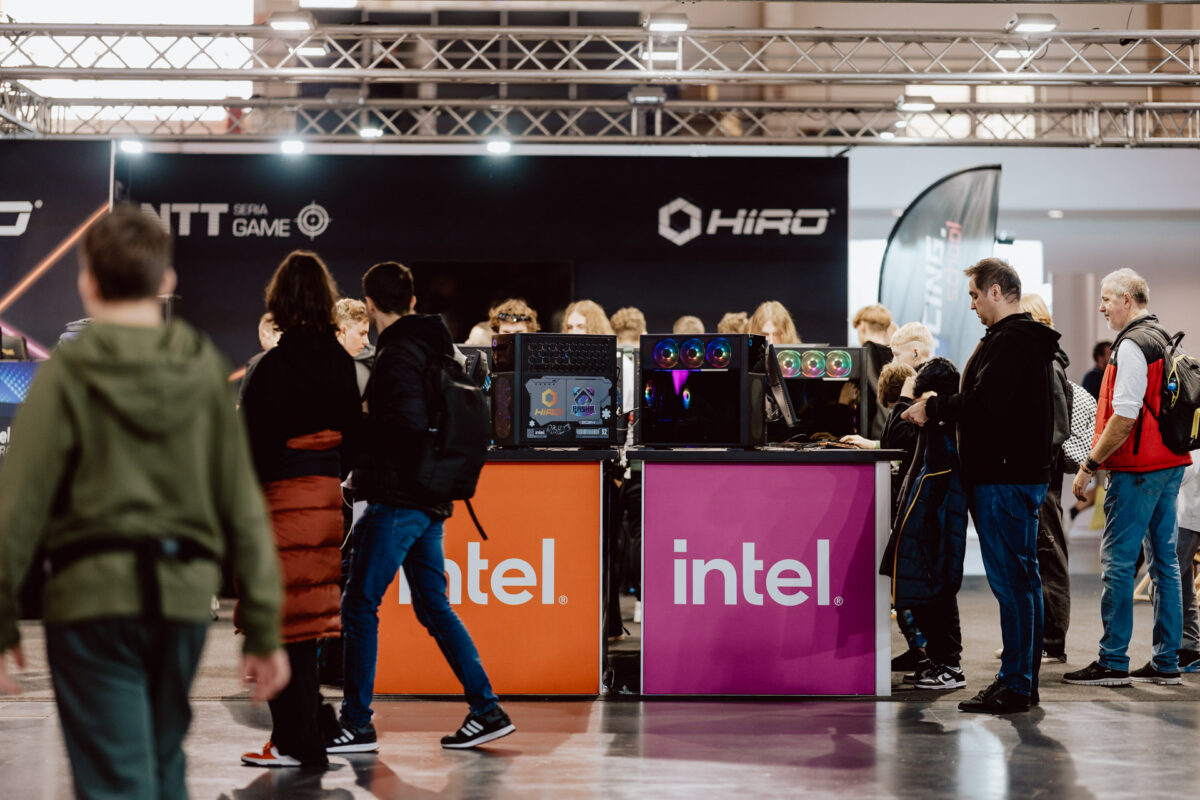 Menschen versammeln sich um die Stände der Marke Intel auf einer Technologieveranstaltung, mit ausgestellten Spielecomputern und verschiedenen Bannern im Hintergrund. Die Atmosphäre ist geschäftig und lebhaft, die Teilnehmer interagieren und erkunden die Exponate. 