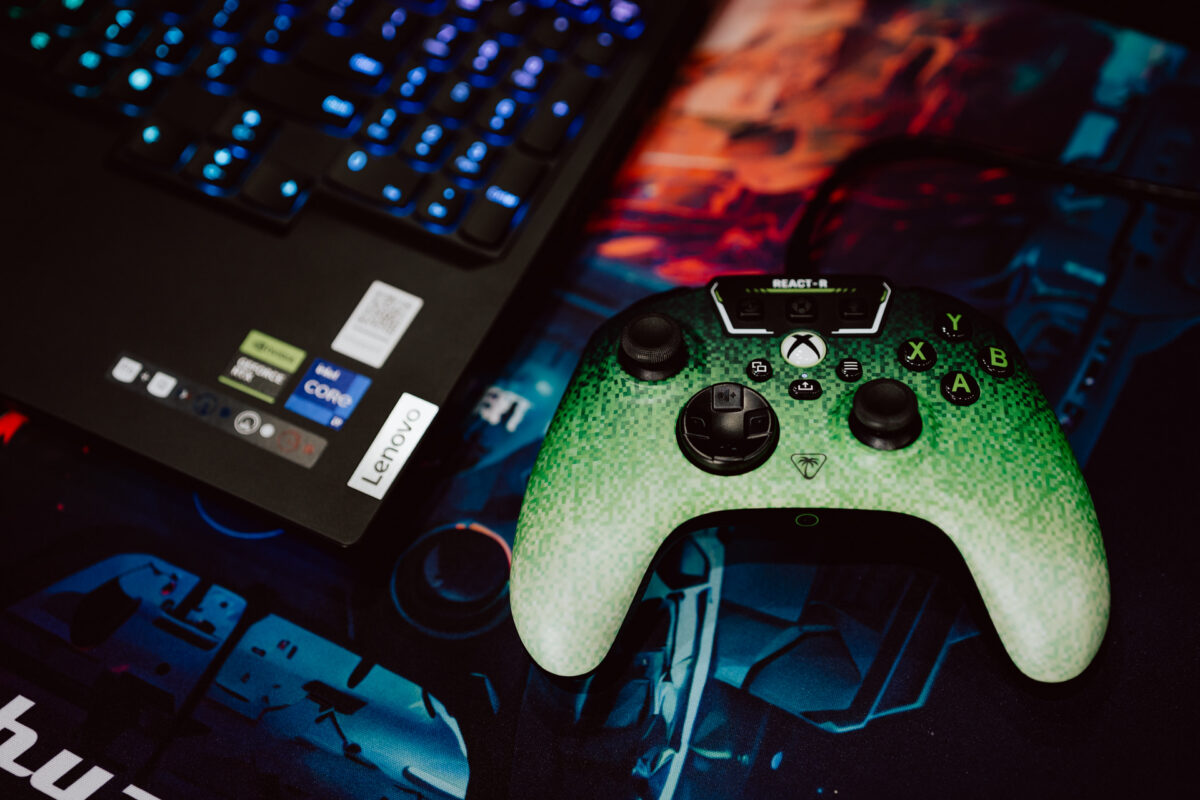 Ein grüner Xbox-Controller liegt auf einem bunten Gaming-Mauspad neben einem aufgeklappten Lenovo-Laptop mit einer blau leuchtenden Tastatur.