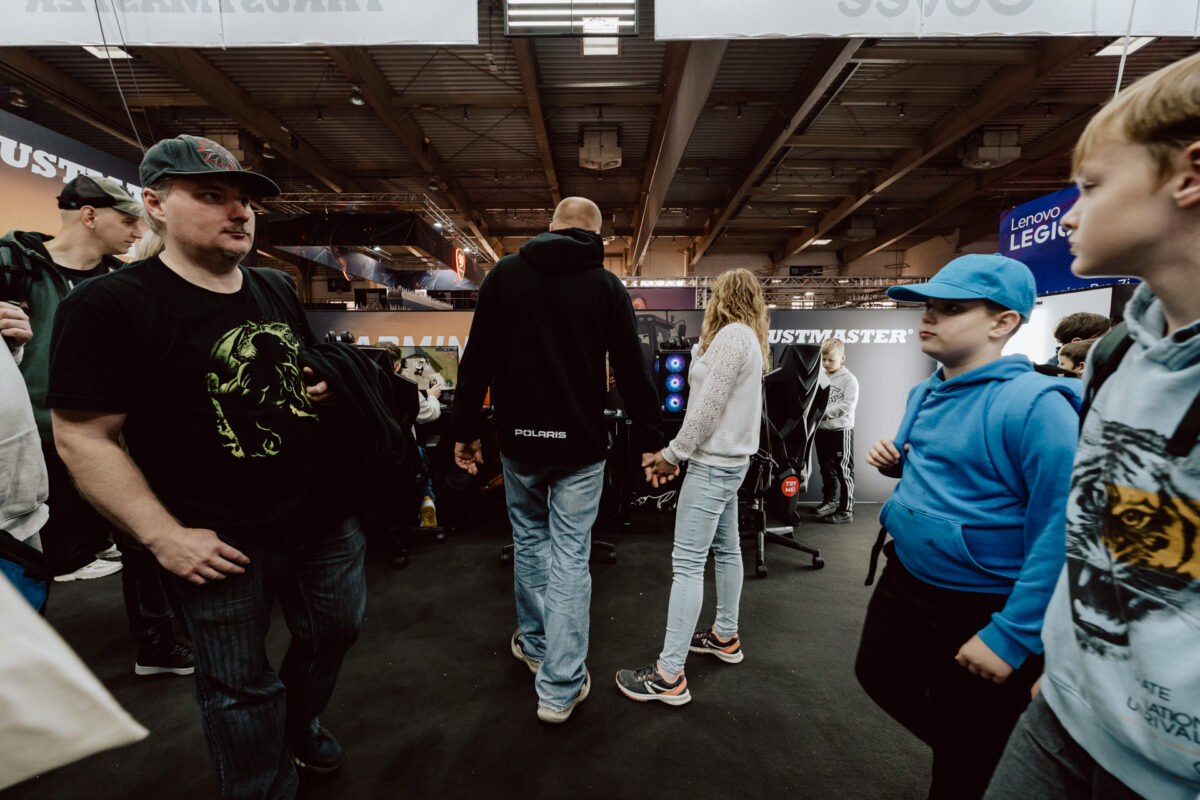 Eine Menschenmenge läuft durch eine geschlossene Veranstaltung oder einen Kongress. Einige tragen Hüte und Kapuzenpullis. Im Hintergrund sind Stände und Banner mit Markennamen zu sehen. Die Atmosphäre ist geschäftig und lebhaft.   