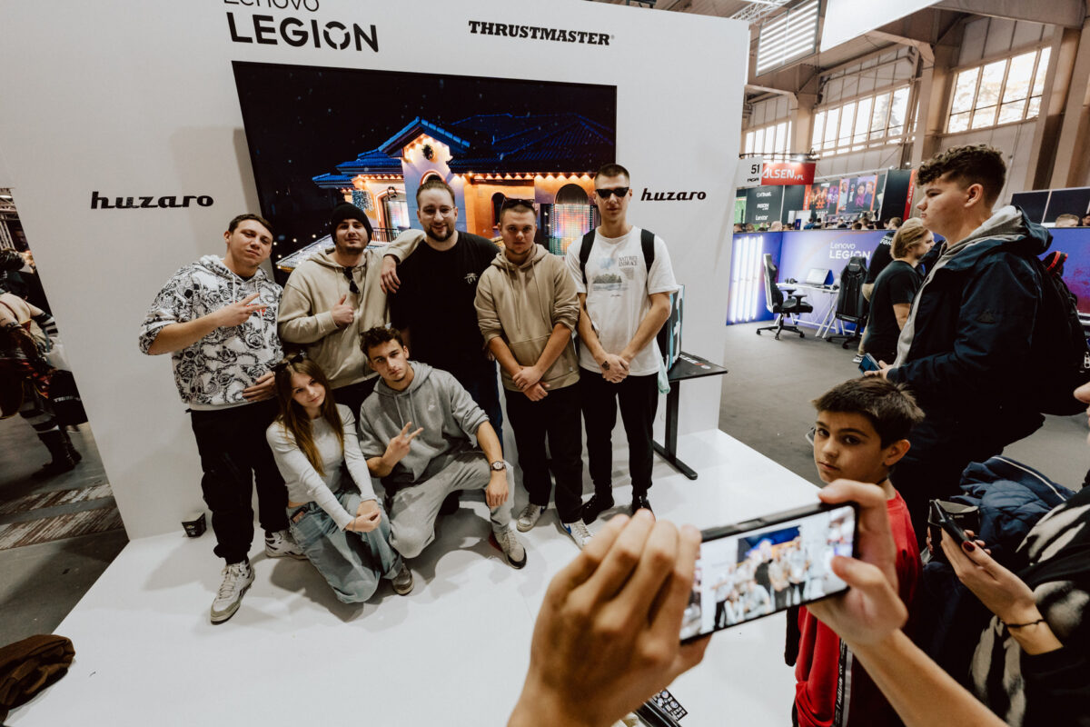 Eine Gruppe junger Leute posiert gemeinsam für ein Foto an einem Stand mit den Marken 'Lenovo Legion', 'Thrustmaster' und 'Huzaro'. Jemand im Vordergrund hält ein Telefon in der Hand und nimmt die Gruppe auf. 