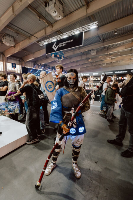 Eine Person in einem detailgetreuen Samurai-Cosplay, die einen langen Stab in der Hand hält und ein blaues Gewand, eine Maske und eine Rüstung trägt, steht in einer überfüllten Kongresshalle, umgeben von Besuchern und Ausstellungsständen.