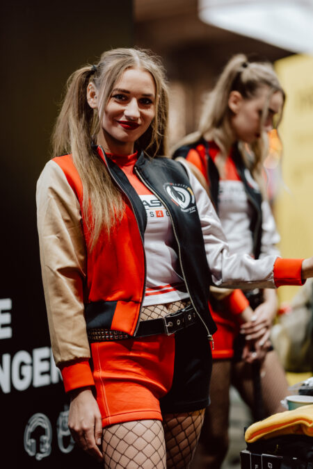 Zwei Frauen in sportlichen, farbenfrohen Outfits mit roten und schwarzen Jacken, Shorts und Cabaret-Strümpfen stehen in einem Raum. Die vordere Frau lächelt in die Kamera, während die andere nach unten schaut, teilweise unscharf. 