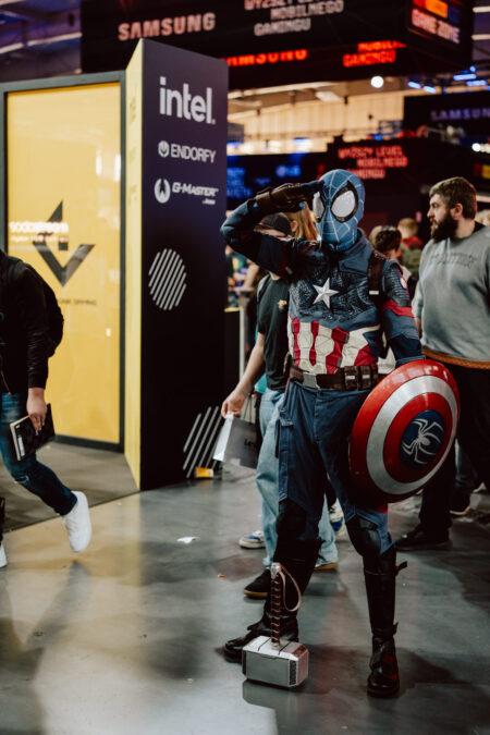 Eine Person in einem Spider-Man- und Captain America-Kostüm posiert auf der Convention mit dem Schild von Captain America und dem Hammer von Thor, während im Hintergrund Stände und Veranstaltungsteilnehmer zu sehen sind.