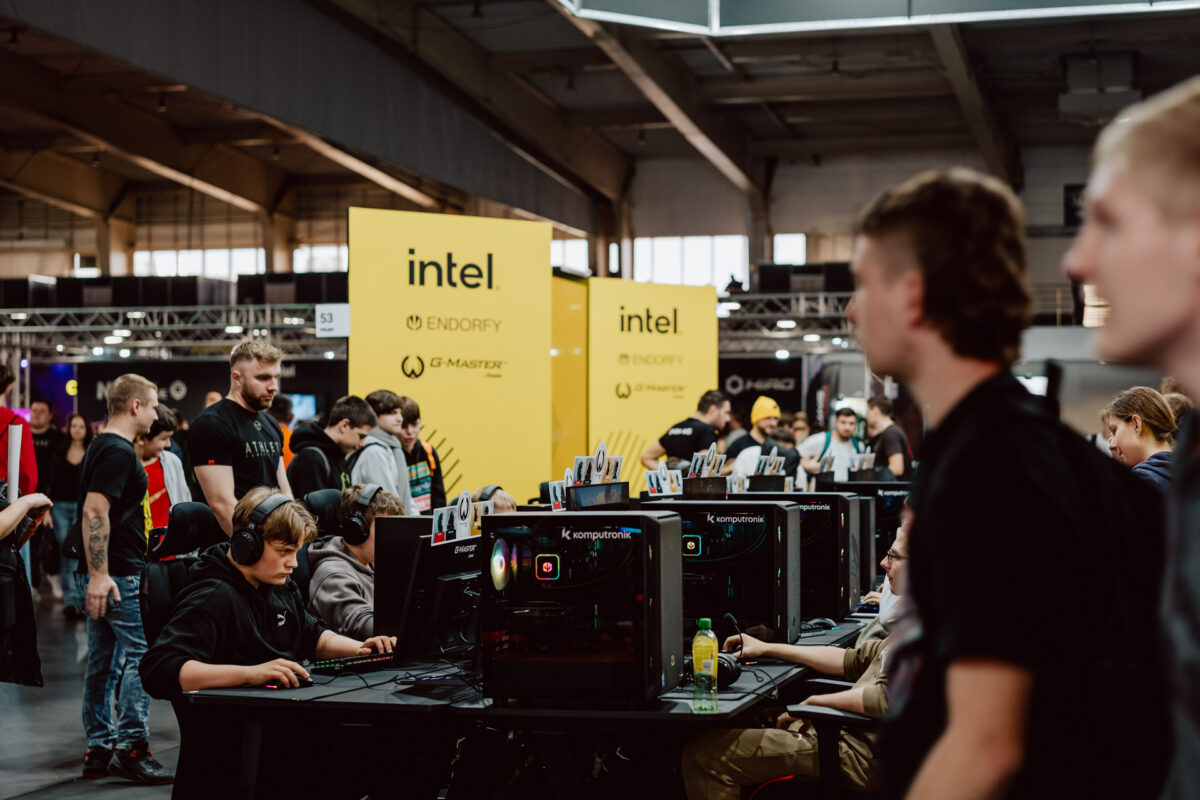 Eine Gruppe von Leuten nimmt an einem Gaming-Turnier auf einer Convention teil. Zuschauer blicken auf sie zurück. Im Hintergrund sind große gelbe Intel-Banner und verschiedene Spielgeräte zu sehen.  