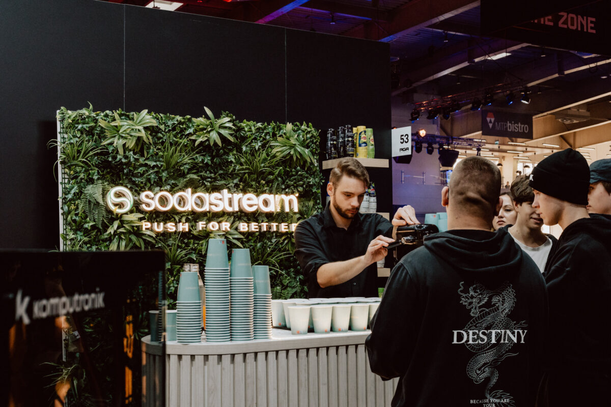 Ein Mann serviert einer Gruppe von Menschen Getränke an einem Sodastream-Stand mit einer grünen Wand und einem Stapel von Bechern in einem belebten Veranstaltungsraum.