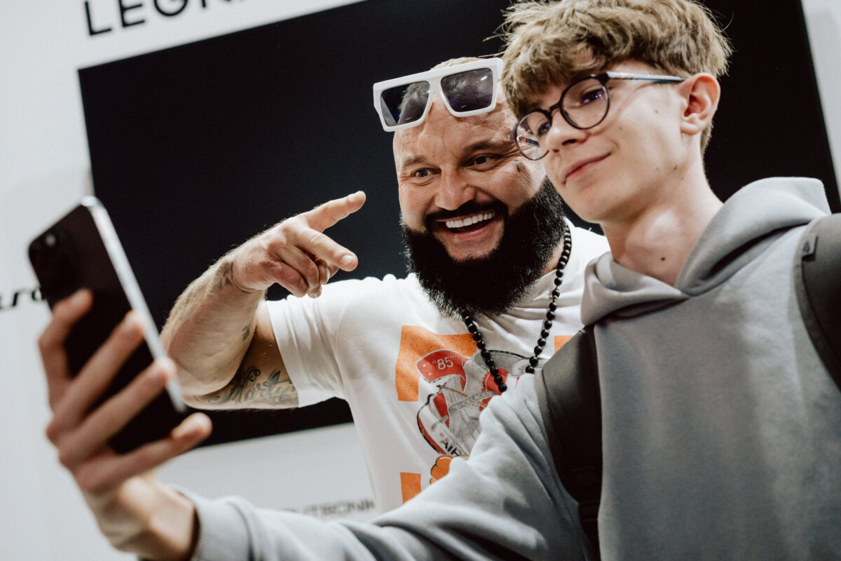 Zwei Personen machen ein Selfie: ein lächelnder bärtiger Mann mit Sonnenbrille auf dem Kopf und ein junger Mann mit Brille und Kapuzenpulli. Beide sehen glücklich aus und posieren für ein Foto.