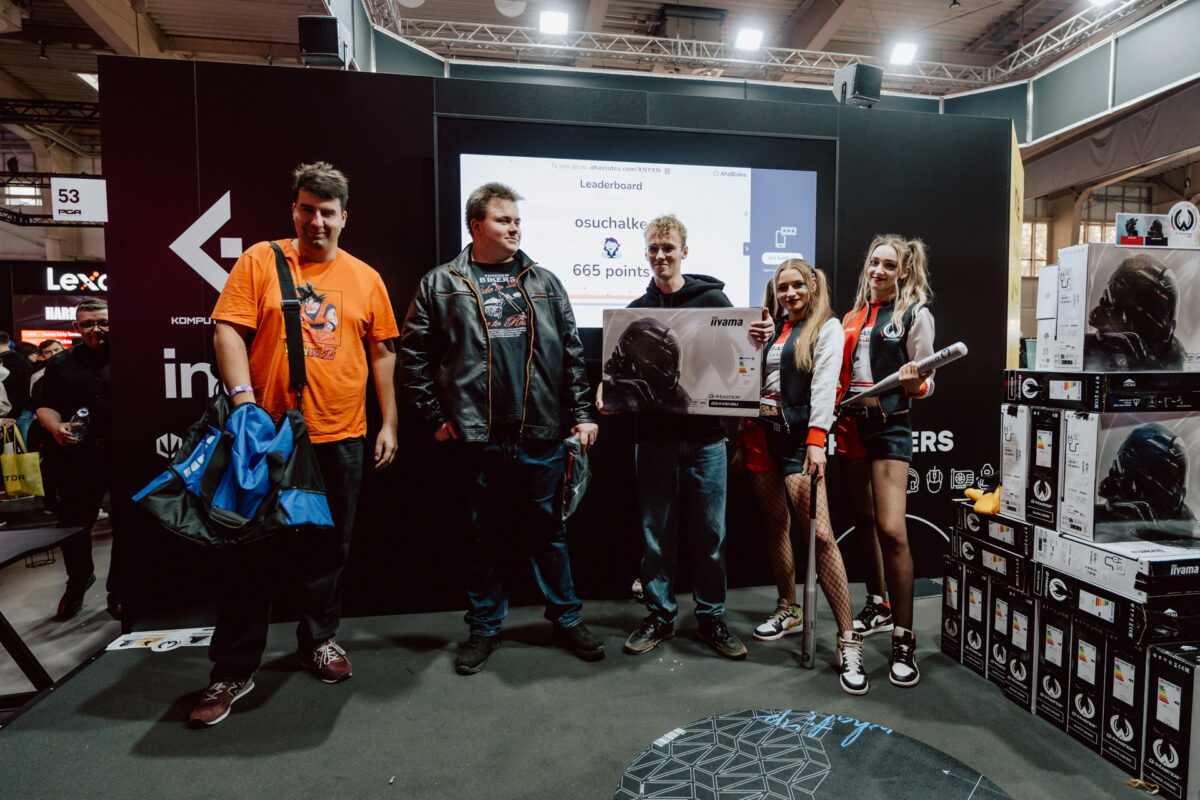 Fünf Personen stehen während der Veranstaltung auf der Bühne; drei halten Preise in der Hand und zwei sind kostümiert. Auf einem großen Bildschirm hinter ihnen ist eine Anzeigetafel zu sehen und rechts davon sind die Boxen gestapelt. Es scheint sich um eine Spiele- oder Technologieveranstaltung zu handeln.  