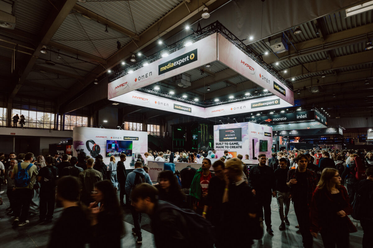 Eine belebte Kongresshalle voller Menschen, von denen viele herumlaufen und einige in der Nähe von Spieleständen und Technologieausstellungen unter hellen Lichtern und großen hängenden Markenbannern stehen.