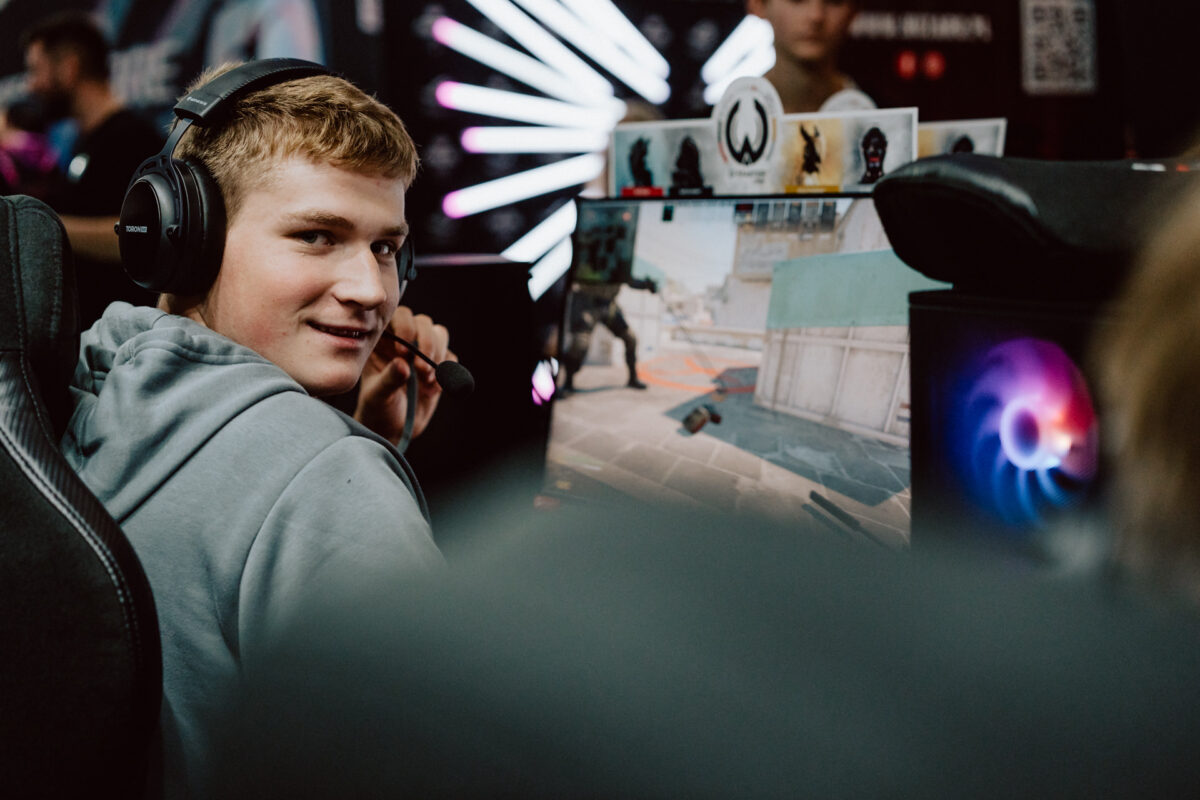 Ein junger Mann mit einem Headset lächelt, während er bei einem Gaming-Event einen Ego-Shooter auf einem Computer spielt. Im Hintergrund sind helle Lichter und andere Spieler zu sehen. 