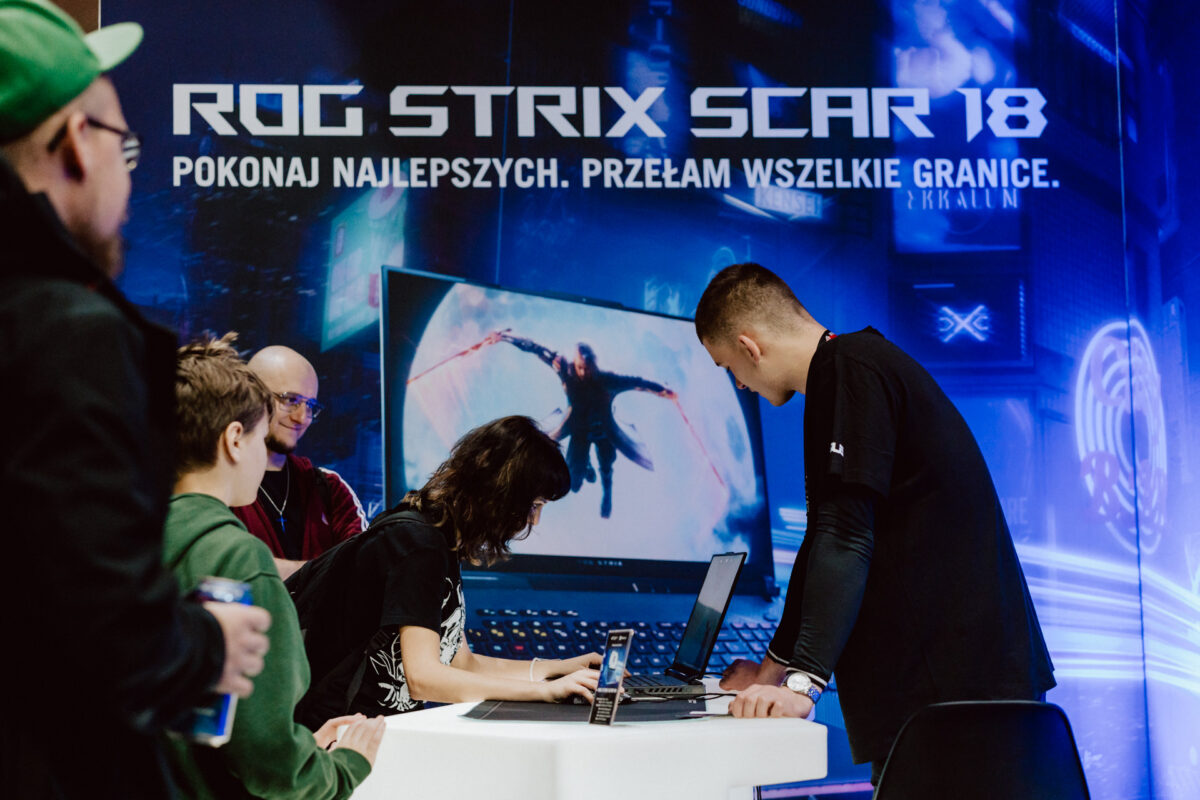 Ein paar Leute versammeln sich um einen Gaming-Laptop am Veranstaltungsstand mit einem großen 'ROG STRIX SCAR 18'-Display und polnischem Text im Hintergrund; auf dem Monitor wird eine digitale Actionszene angezeigt.