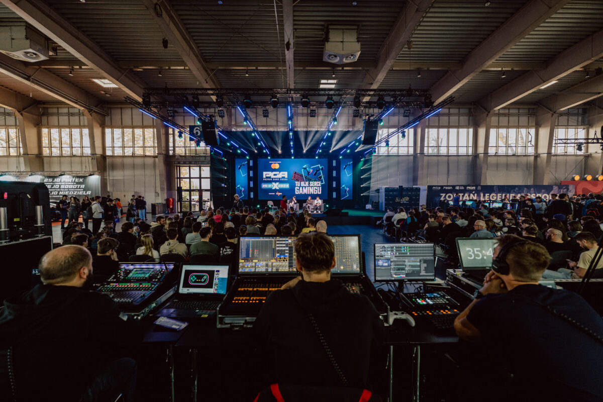 Eine große Veranstaltung in einem Raum, in dem das Publikum gegenüber einer hell erleuchteten Bühne sitzt. Ton- und Lichttechniker bedienen die Geräte in einer Kontrollkabine im Vordergrund. Mehrere Bildschirme und LED-Lichter erhellen die Bühne.  