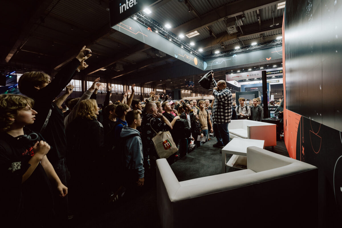 Ein Mann steht auf der Bühne und hält einen Gegenstand für eine Menge aufgeregter Menschen bei einer Indoor-Veranstaltung. Das meist junge Publikum streckt ihm die Hand entgegen. Moderne Beleuchtung und Stände füllen den überfüllten Ausstellungsraum.  