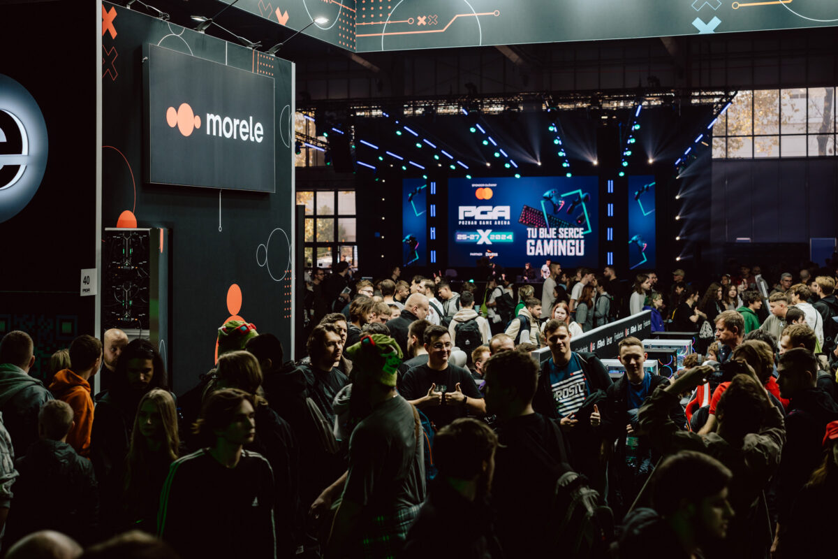 Eine große Menschenmenge versammelt sich zu einer Indoor-Gaming-Veranstaltung mit beleuchteten Schildern, Ständen und einer Bühne mit Live-Bildschirmen und spielbezogenen Bannern im Hintergrund.