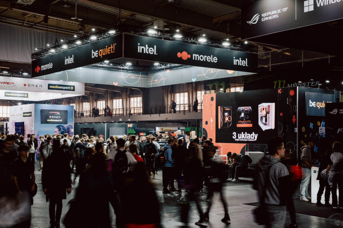 Eine überfüllte Technologie-Messe mit vielen Menschen, die um große Stände von Marken wie Intel, Morele und be quiet! herumlaufen, mit hellen Schildern, digitalen Displays und Spieleinrichtungen in dramatischer Beleuchtung.
