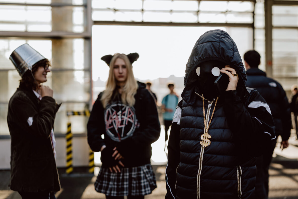 Drei kostümierte Personen stehen in einem Raum. Eine trägt einen eimerförmigen Helm, eine andere hat Katzenohren-Stirnbänder und einen karierten Rock und die dritte hat eine Kapuzenjacke, eine Maske mit runden Augen und eine Kette und hält sich ein Telefon ans Ohr. 