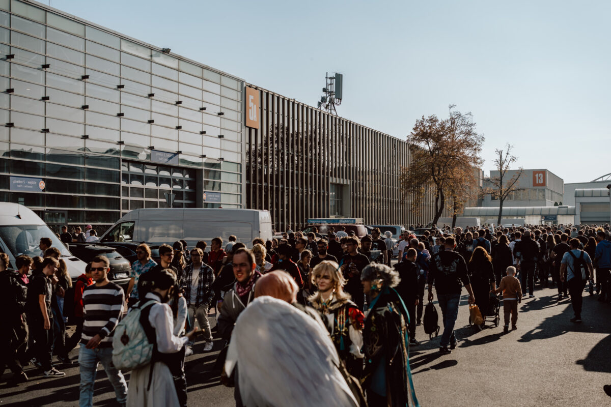 Eine große Menschenmenge, einige in Kostümen, läuft an einem sonnigen Tag vor einem modernen Kongresszentrum mit Glas- und Metallwänden. In der Nähe sind mehrere Lieferwagen geparkt und rechts ist ein Baum zu sehen. 