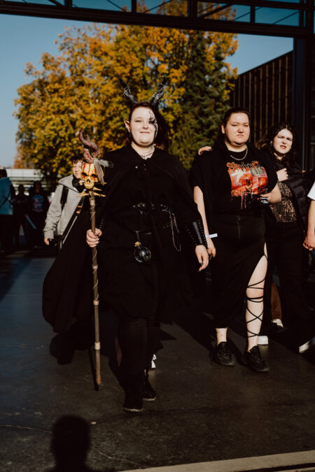 Eine Person in einem schwarzen Fantasiekostüm mit Hörnern und einem Spazierstock geht mit anderen hinein; das Sonnenlicht beleuchtet die Herbstbäume draußen im Hintergrund.
