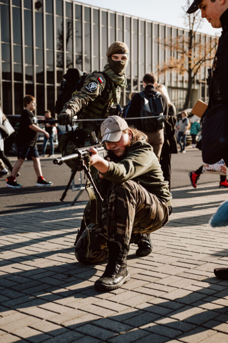 Zwei Personen in militärischer Tarnkleidung, eine hockt und zielt mit einem nachgebauten Gewehr, die andere steht mit verdecktem Gesicht dahinter und zielt. Im Hintergrund gehen Menschen in der Nähe eines großen Glasgebäudes spazieren. 