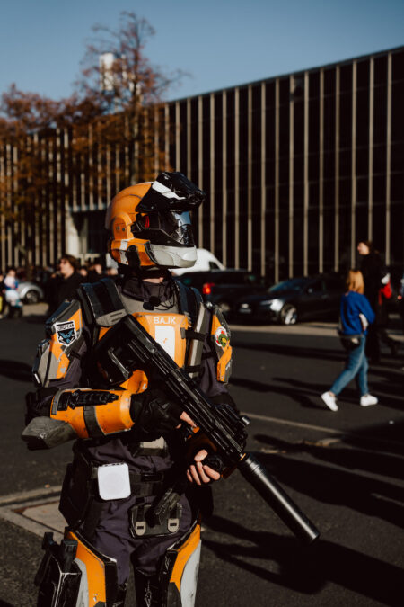 Eine Person in einem orange-schwarzen futuristischen Panzerkostüm hält eine Pistole in der Hand, während sie draußen auf einer Straße steht; im Hintergrund sind Menschen und Autos zu sehen.