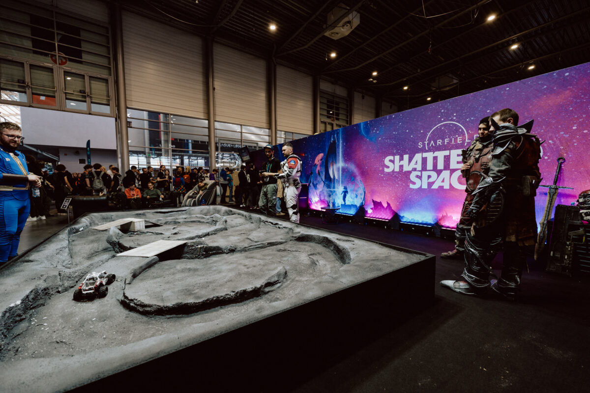 Ein als Astronaut verkleideter Cosplayer steht vor einer Sci-Fi-Bühne mit dem Hintergrund von 'Starfield: Shattered Space', während die Menge ein Miniatur-Felsgebiet mit einem kleinen Rover beobachtet.
