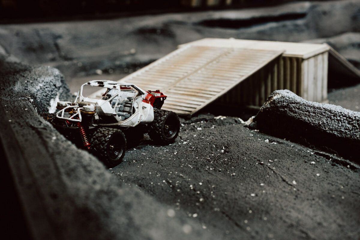 Ein kleines Modell eines geländegängigen Fahrzeugs, das einem Rover ähnelt, ist auf einem unebenen, dunklen Boden mit verstreuten Trümmern geparkt. Im Hintergrund ist eine hölzerne Rampe oder Brückenstruktur zu sehen. 