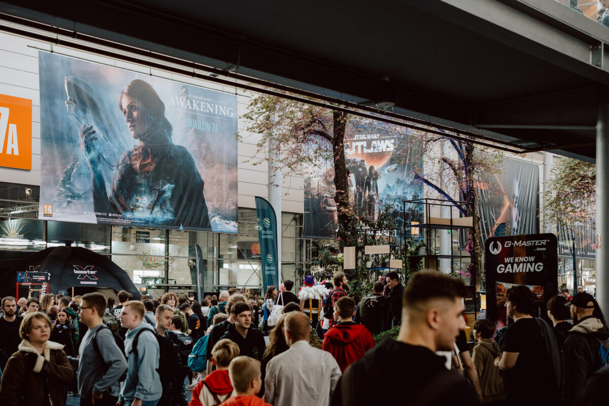 Eine große Menschenmenge versammelt sich auf einem Spielekongress. Spieleposter, darunter eines für 'AWAKENING', hängen über der Veranstaltung. Stände, Banner und Spieleauslagen füllen den überfüllten Innenraum, in dem viele Teilnehmer herumlaufen und Kontakte knüpfen.  