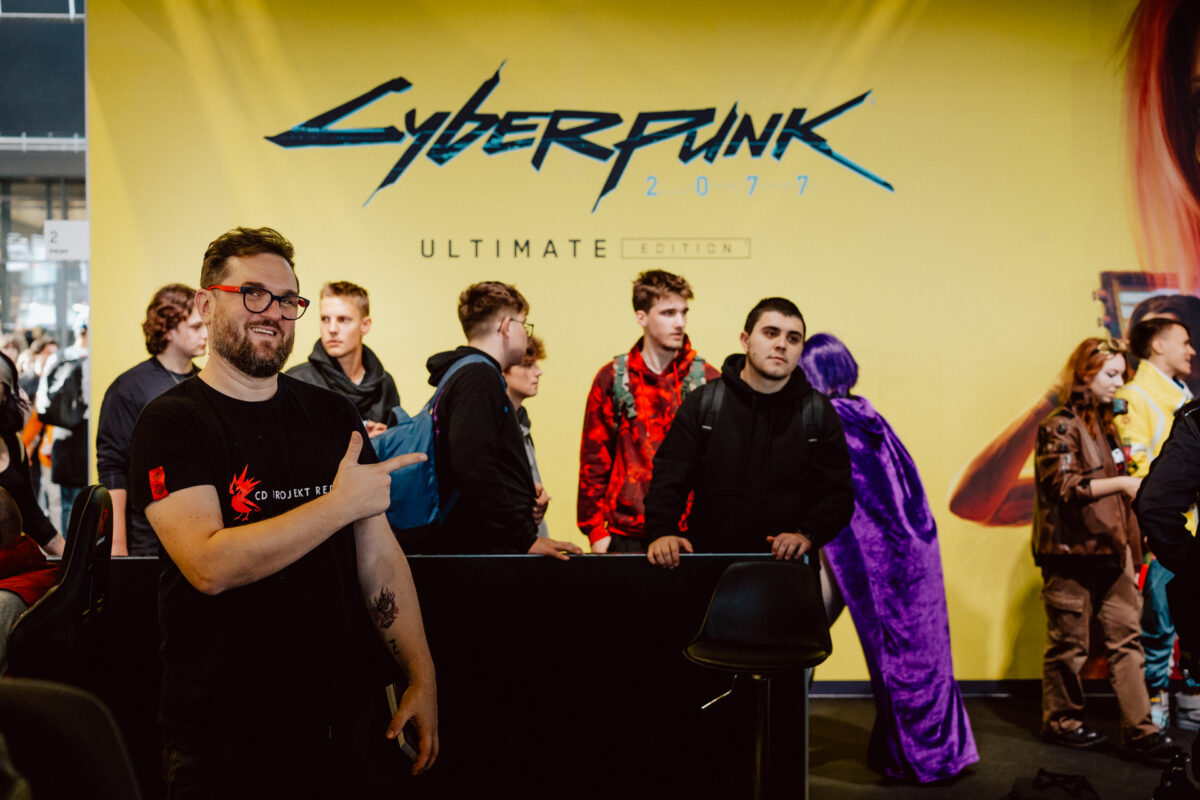 Der Mann lächelt und zeigt auf eine große gelbe Anzeige von 'Cyberpunk 2077 Ultimate Edition', während eine Menschenmenge vor einem Stand auf der Veranstaltung steht und wartet.