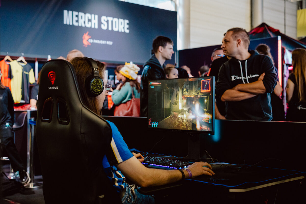 Eine Person, die Kopfhörer trägt, spielt während der Veranstaltung ein Videospiel auf einem Computer, während im Hintergrund Menschen in der Nähe eines Verkaufsstandes stehen und sich unterhalten.