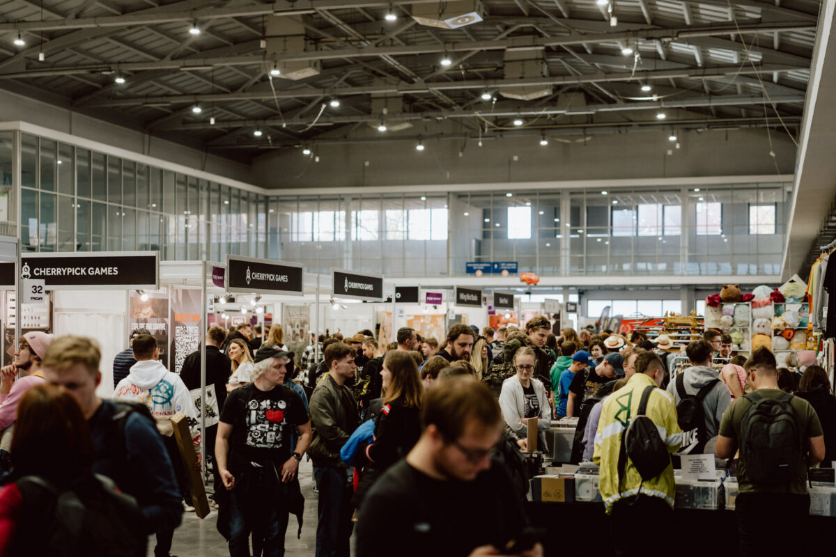 Eine große Menschenmenge läuft durch das überdachte Kongresszentrum, das mit Verkaufsständen und Schildern gefüllt ist, darunter eines für 'Cherrypick Games'. Helle Lichter erhellen die geschäftige, geräumige Halle. 