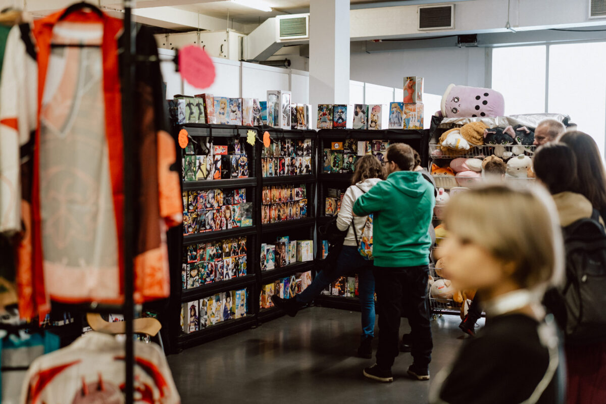 Menschen stöbern in den Manga- und Anime-Artikeln, die in den Regalen auf der Convention oder im Geschäft ausgestellt sind. Im Vordergrund sind Cosplay-Kostüme zu sehen, während im Hintergrund Plüschtiere und verschiedene Sammlerstücke zu sehen sind. 