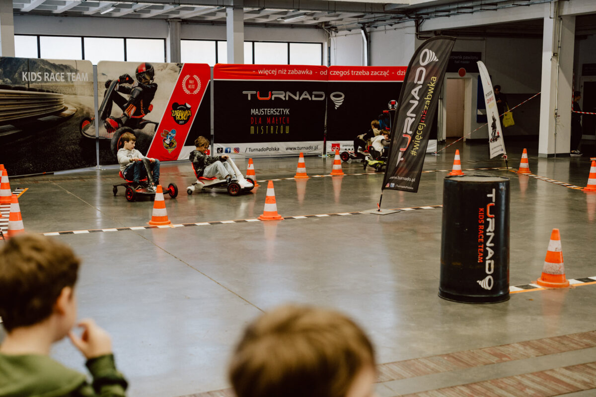 Kinder fahren Gokarts auf einer mit orangefarbenen Kegeln markierten Indoor-Rennstrecke. Im Hintergrund sind Banner und Werbeschilder für die Rennveranstaltung zu sehen, während ein paar Leute vom Rand aus zusehen. 