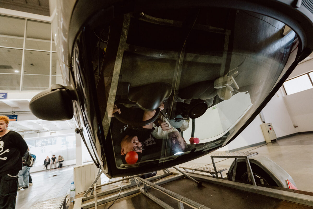 Ein umgedrehtes Auto auf einer Plattform in einem Raum, in dem zwei Personen sitzen, die lächeln und die Sitze festhalten. Schaulustige beobachten die Situation im Hintergrund, in der Nähe der großen Fenster und des Betonbodens. 