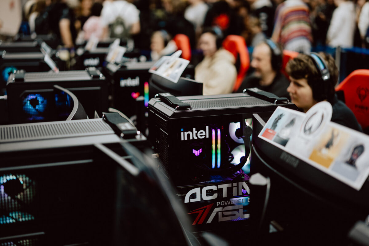 Eine Reihe von Gaming-Computern und Monitoren mit Spielern, die an jeder Station Headsets tragen und an einem Gaming-Event teilnehmen, mit einer unscharfen Menschenmenge im Hintergrund.